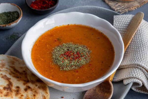 Tarhana Soup Recipe – Give Recipe A bowl of tarhana soup garnished with dried mint and red pepper flakes, some bread and a wooden spoon on the side.