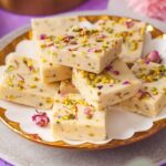 A plate stacked with pieces of creamy white thandai fudge topped with chopped pistachios and rose petals
