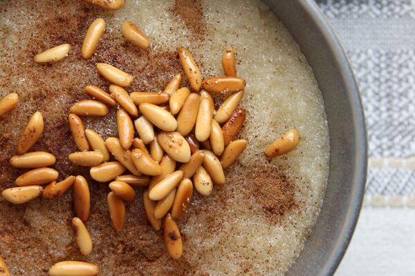 Toasted Semolina Pudding (Mamounieh) with Pine Nuts & Cinnamon Close up, cut off shot of a grey bowl with Mamounieh a semolina pudding topped with toasted pine nuts and cinnamon with a grey cloth napkin next to the bowl