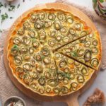 A round pizza topped with spiral-cut zucchini slices is served on a wooden board, with one slice slightly separated from the rest. A bowl of cheese and a pepper mill are nearby.