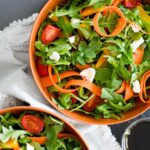 arugula salad in orange round bowls with cherry tomatoes, crumbled goat cheese, ribbon carrots, and bell peppers.