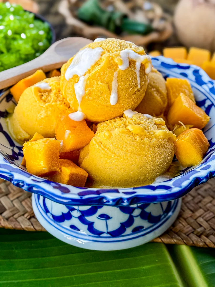 Asian mango ice cream served with fresh mango chunks and a drizzle of coconut cream in a traditional Thai porcelain dish.