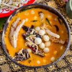 A close-up of Thai chicken feet curry in a coconut shell bowl, highlighting the creamy red curry broth and fresh vegetables.