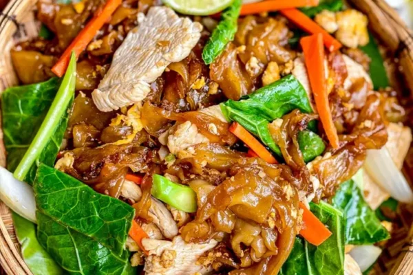 Chicken Pad See Ew (Thai Stir Fried Noodles) Chicken pad see ew with wide rice noodles, leafy greens, carrots, and garlic on a decorative fabric.