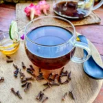 A clear glass cup of clove tea on a wooden platter surrounded by whole cloves, honey, and a slice of lime, with a teapot in the background.