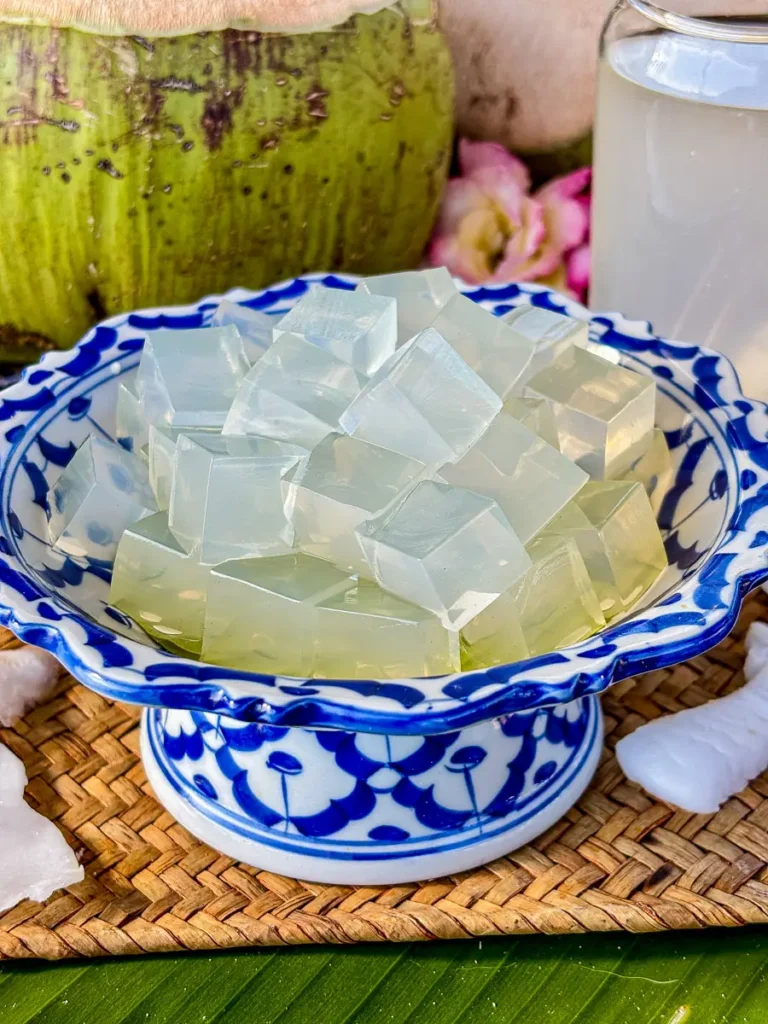 Close-up of coconut jelly cubes in a traditional Thai porcelain bowl, with fresh coconut water and coconut pieces nearby.