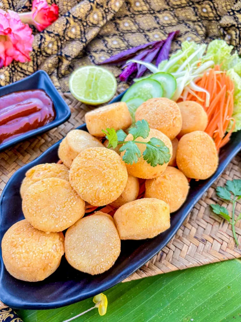 Best Crispy Fried Egg Tofu Recipe Crispy fried egg tofu pieces served with shredded vegetables, cucumber, and dipping sauce on a black plate over a woven mat and banana leaf.