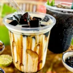 Close-up of two drinks; a black grass jelly milk drink with jelly cubes, and a dark black grass jelly drink with ice. Ingredients like brown sugar, lime, and jelly cubes are arranged around.