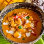 Thai mango chicken curry in a coconut bowl garnished with fresh cilantro and red chili, with a fresh mango in the background.