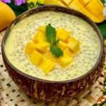 Mango sago dessert served in a coconut shell, topped with mango cubes and a mint leaf.