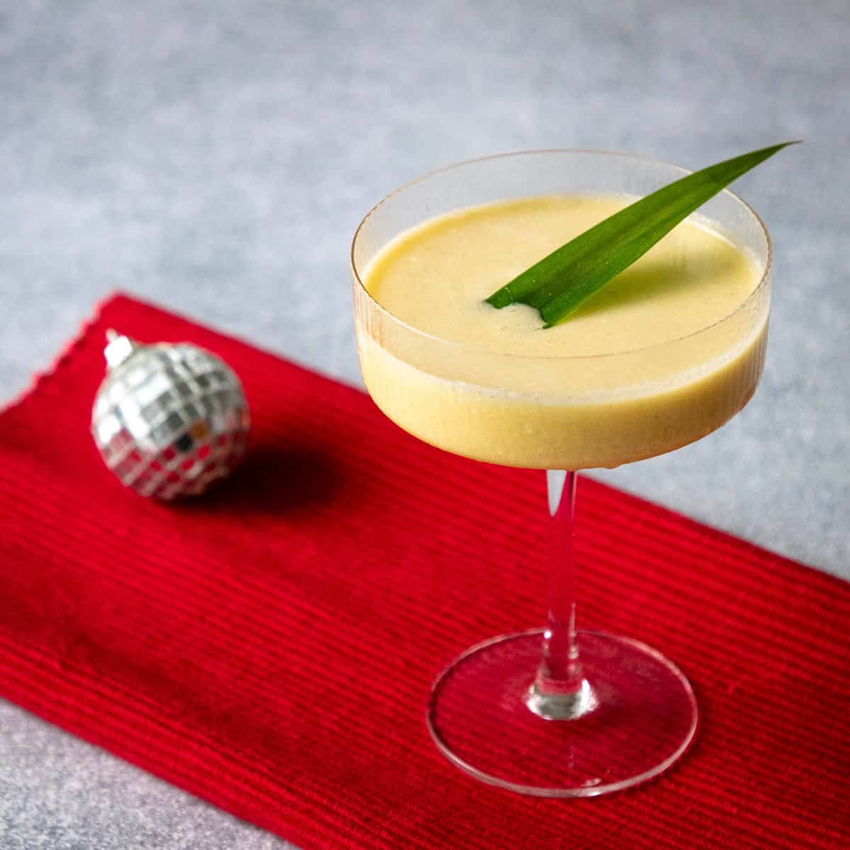Thai Mango Sticky Rice Cocktail mango sticky rice cocktail in a coupe glass with a pandan leaf garnish and a small disco ball in the background.