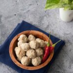 a bowl of moo deng meatballs with 2 chilies and a plant in the background
