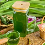Glass jug of green pandan syrup with a small glass cup, surrounded by pandan leaves and decorative flowers on a woven tray.