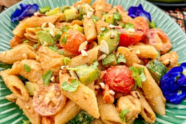 Thai peanut chicken pasta salad with penne, cucumber, cherry tomatoes, and chopped peanuts served in a green dish garnished with butterfly pea flowers.