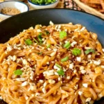 Thai peanut noodles garnished with peanuts, cilantro, and green onions in a black dish, with small bowls of ingredients in the background.