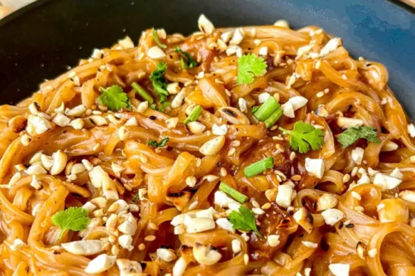 Thai peanut noodles garnished with peanuts, cilantro, and green onions in a black dish, with small bowls of ingredients in the background.