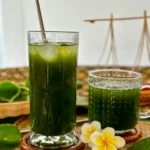 Glass of pennywort drink with a stainless steel straw, next to a smaller mug, tropical flowers, and fresh pennywort leaves.