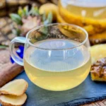 Pineapple tea on a slate tray with fresh ginger slices, a cinnamon stick, pineapple skin, and a teapot in the background.