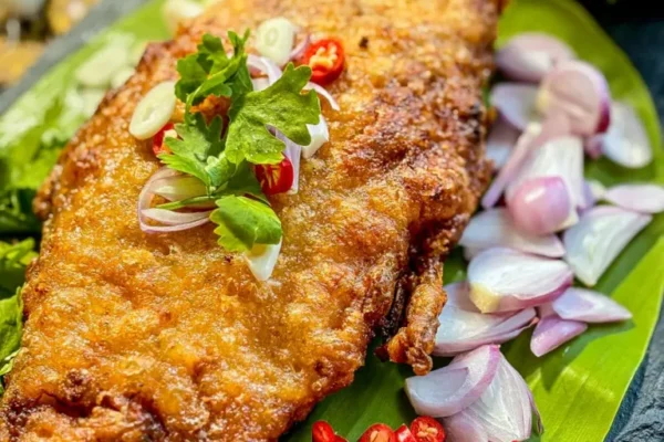 Whole Thai pla som fish deep-fried until golden, garnished with red chili, shallots, lime, and coriander on a green banana leaf.