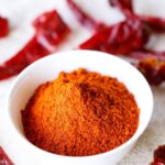 red chilli powder in a white bowl with dried red chillies on top and at the bottom of the bowl on a jute cloth.