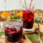 Pouring vibrant red roselle juice into a glass over ice, surrounded by dried roselle buds and Thai décor.