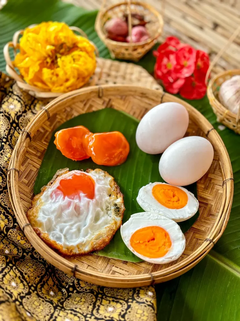 How to Make Salted Egg (Kai Kem) 1 Thai salted eggs shown raw, fried, and boiled, displayed on banana leaf in a woven basket with decorative flowers.