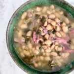 A green pottery bowl filled with smoky white bean soup showing bits of ham and kale in a mostly clear broth