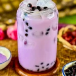 Iced taro milk tea with black boba pearls in a clear glass jar, served over ice on a woven mat with taro powder and tapioca pearls on the side.