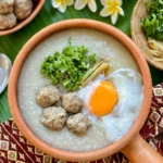 Traditional Thai congee with minced pork, poached egg, ginger, green onion, and coriander in a rustic clay pot.