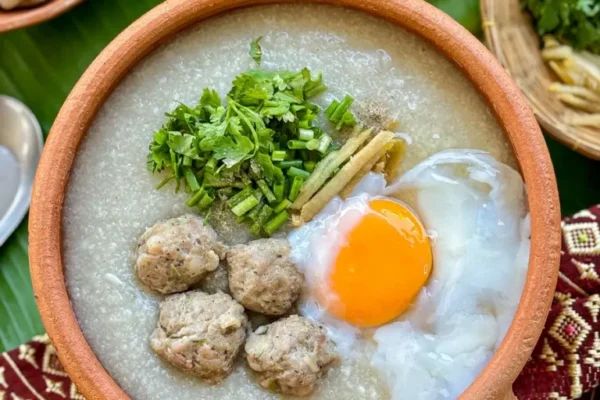 Traditional Thai congee with minced pork, poached egg, ginger, green onion, and coriander in a rustic clay pot.