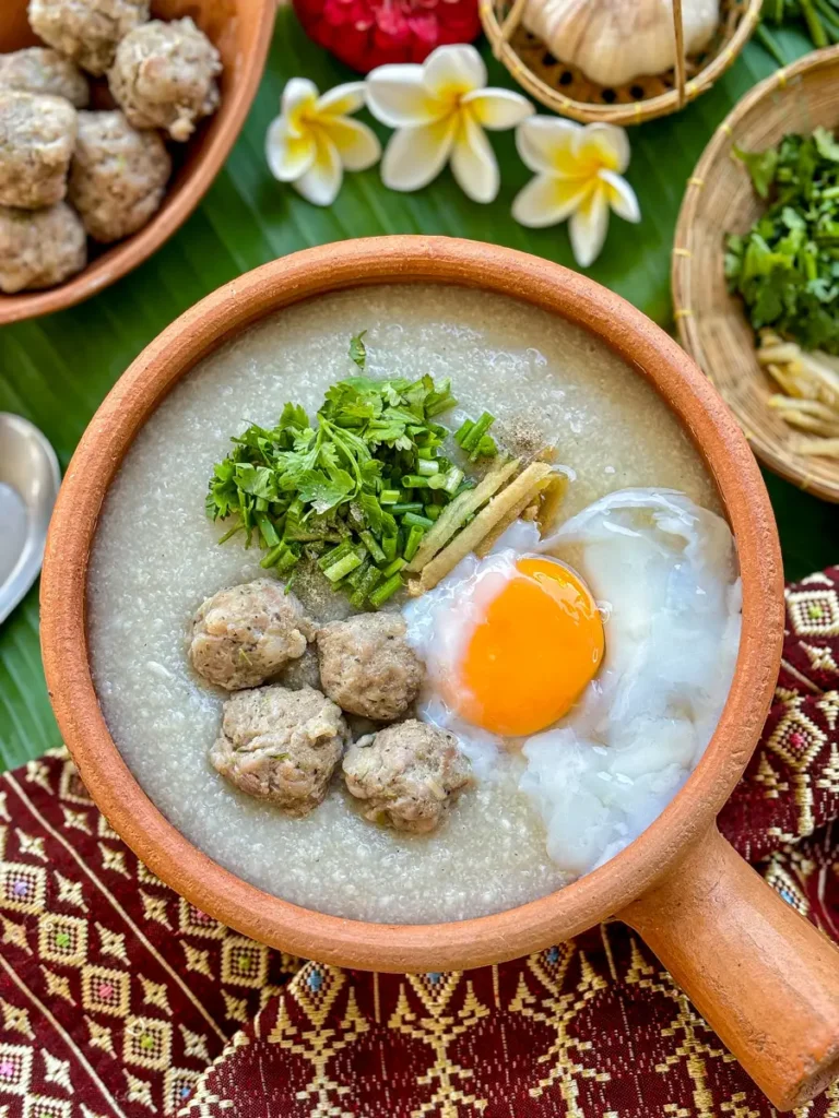 Traditional Thai congee with minced pork, poached egg, ginger, green onion, and coriander in a rustic clay pot.