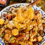 Close-up of Thai cry baby noodles in a blue patterned bowl, featuring golden stir-fried noodles, tofu, sausage, and fresh herbs.