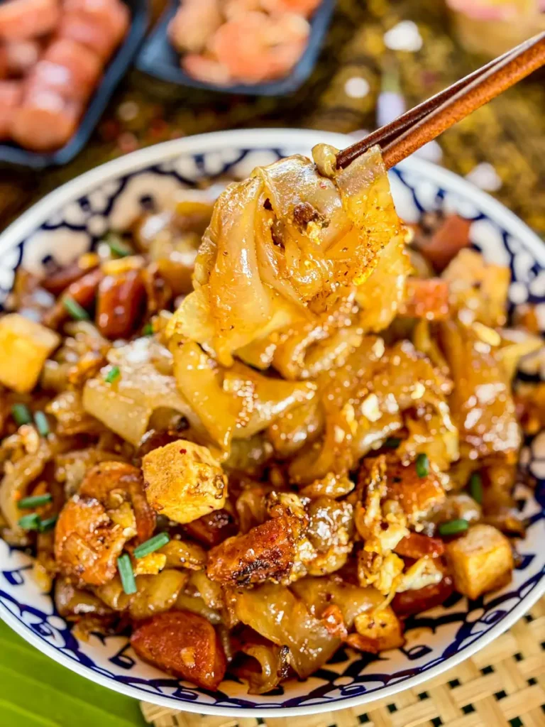 Close-up of Thai cry baby noodles in a blue patterned bowl, featuring golden stir-fried noodles, tofu, sausage, and fresh herbs.