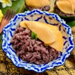 Thai sankaya custard over black sticky rice served in banana leaf cups with mango in the background.