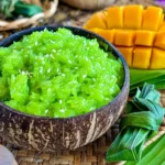 Thai sweet sticky rice in a coconut bowl, glowing green and topped with sesame seeds, paired with cubed mango slices and a decorative pandan bundle on a rustic mat.