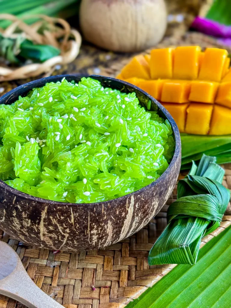 Thai sweet sticky rice in a coconut bowl, glowing green and topped with sesame seeds, paired with cubed mango slices and a decorative pandan bundle on a rustic mat.