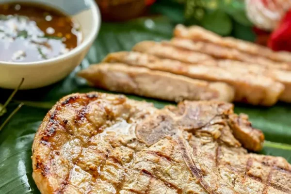 Grilled Thai pork chop served on banana leaf with sticky rice and dipping sauce, beautifully charred and juicy.