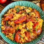 A bowl of Thai tomato and egg stir-fry garnished with green onions, served alongside a bowl of rice, fresh tomatoes, and eggs.