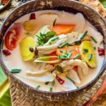A bowl of creamy vegan Thai mushroom soup with coconut milk and vegetables, served in a coconut shell on a woven mat with garnishes.