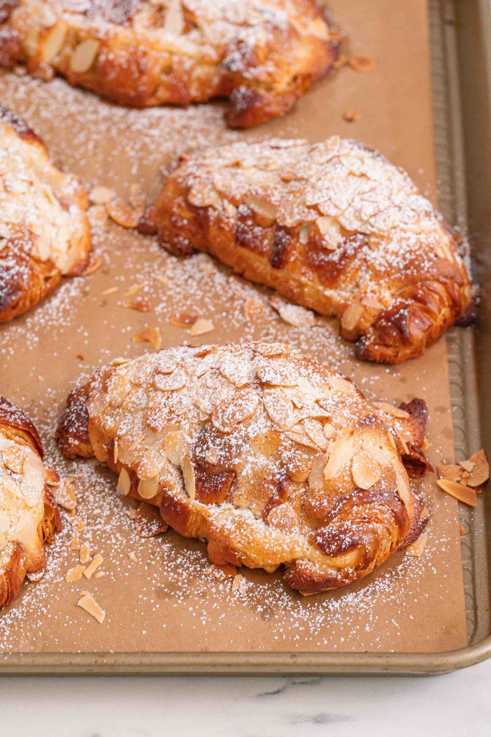 Three almond croissants, topped with sliced almonds and powdered sugar, are arranged on a baking sheet lined with parchment paper.