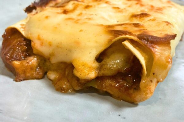 A slice of beef cannelloni seen from the side on a light grey plate with melted cheese coming out from the side.