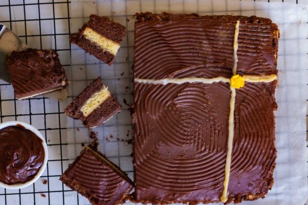 Bombay bakery chocolate cake (Copycat Recipe) A top down shot of a chocolate vanilla cake. The cake is cut in half, and placed on parchment paper on top of a baking rack. There are 4 cake slices along with a bowl of chocolate ganache placed in front of the cake.