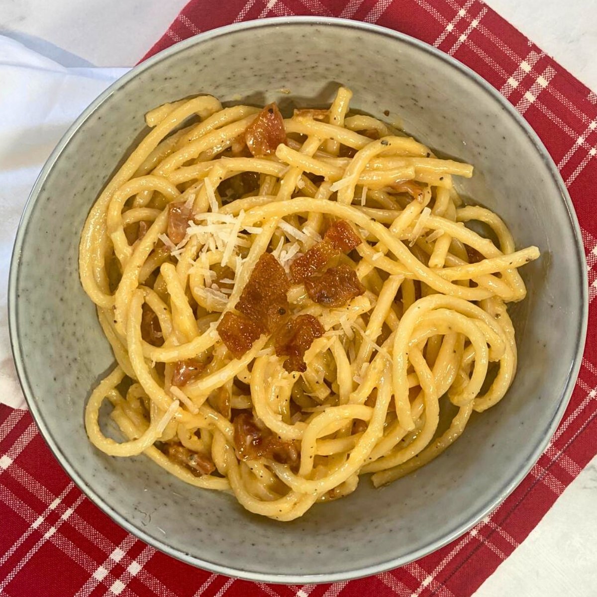 Grey bowl filled with bucatini carbonara with freshly grated cheese and crispy guanciale on top.
