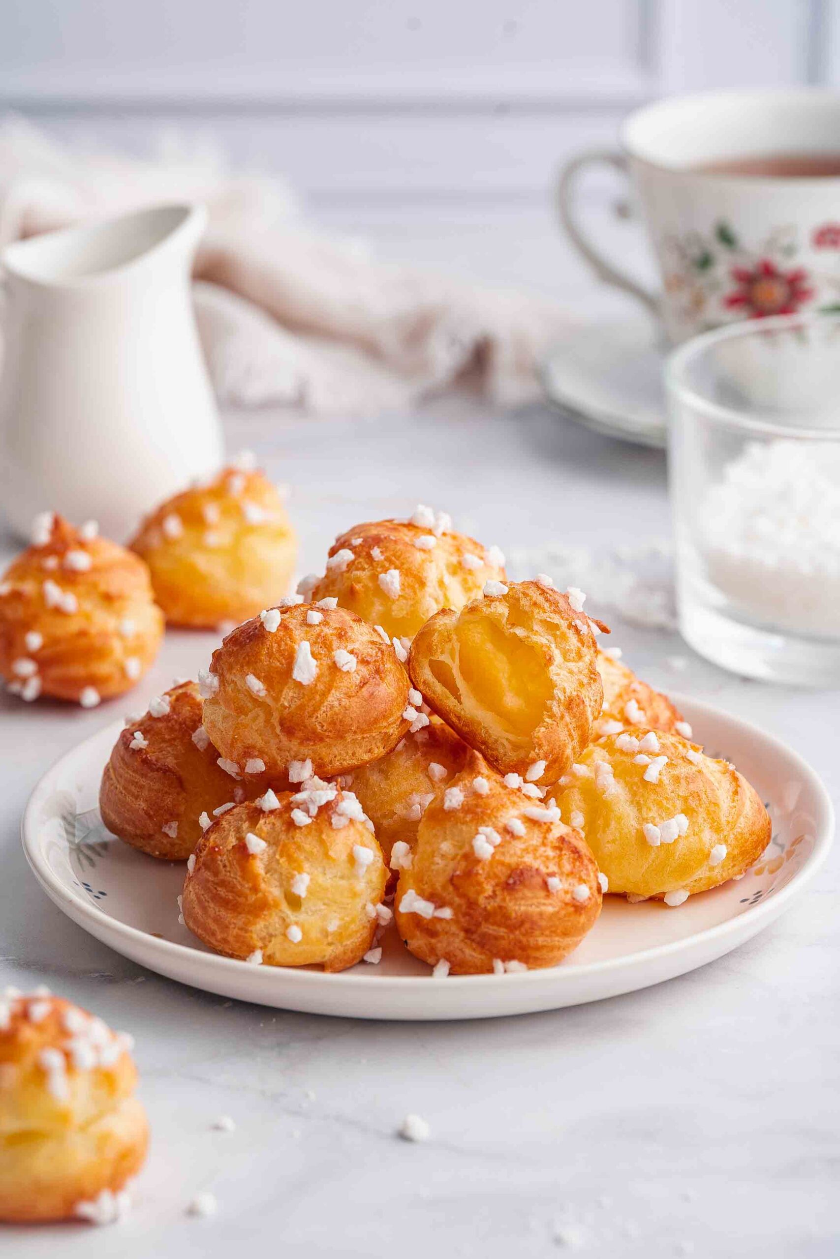 Chouquettes (French Sugar Puffs) | Géraldine's delicacies 1 A plate of golden chouquettes pastries topped with pearl sugar, surrounded by a cup, glass and small pitcher on a light-colored table.