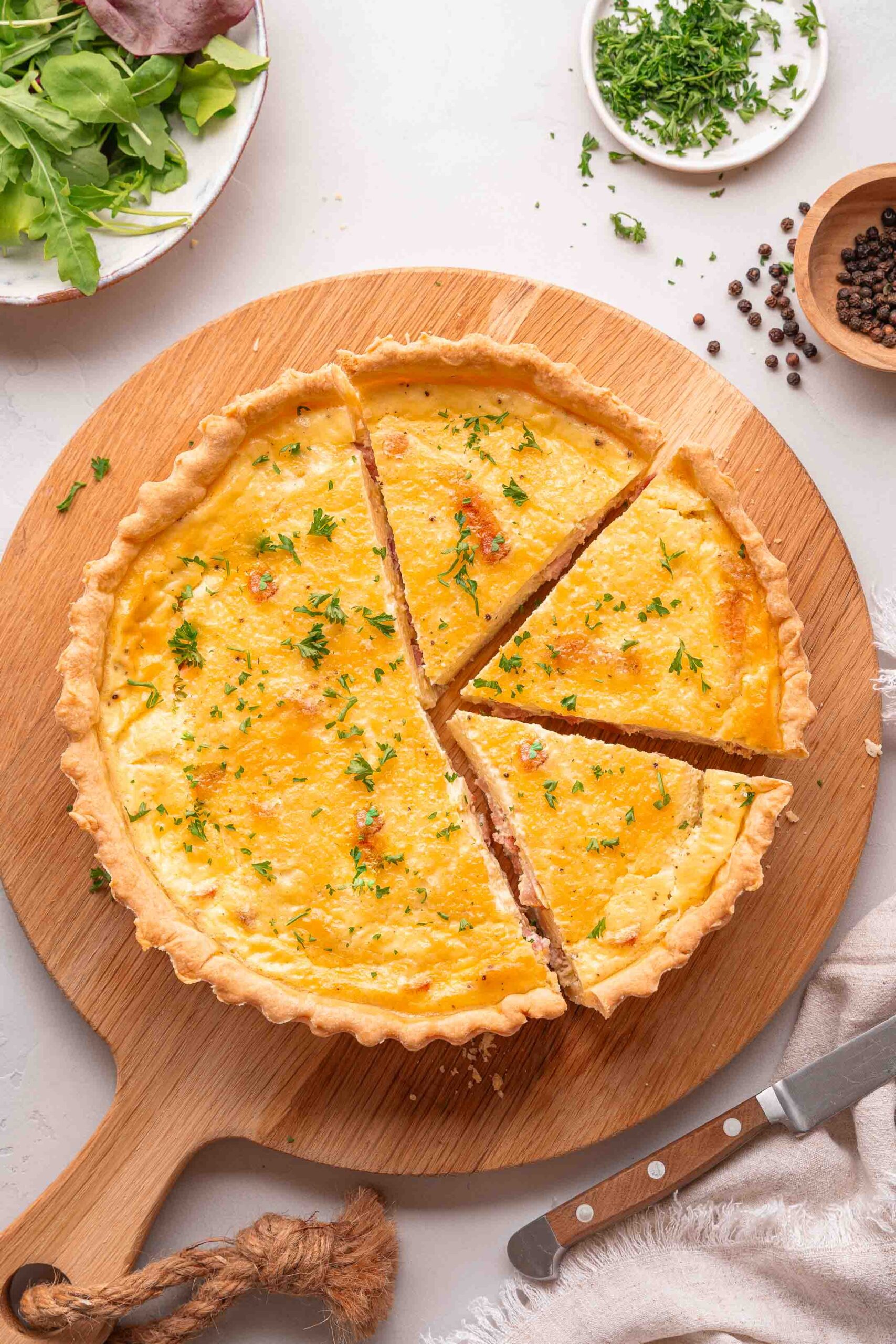 A baked quiche Lorraine with four slices cut, garnished with chopped herbs, sits on a wooden serving board next to a knife, greens, and small bowls of pepper and herbs.