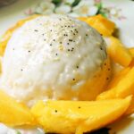 A creamy rice dessert topped with sesame seeds, surrounded by fresh mango slices on a decorative plate.