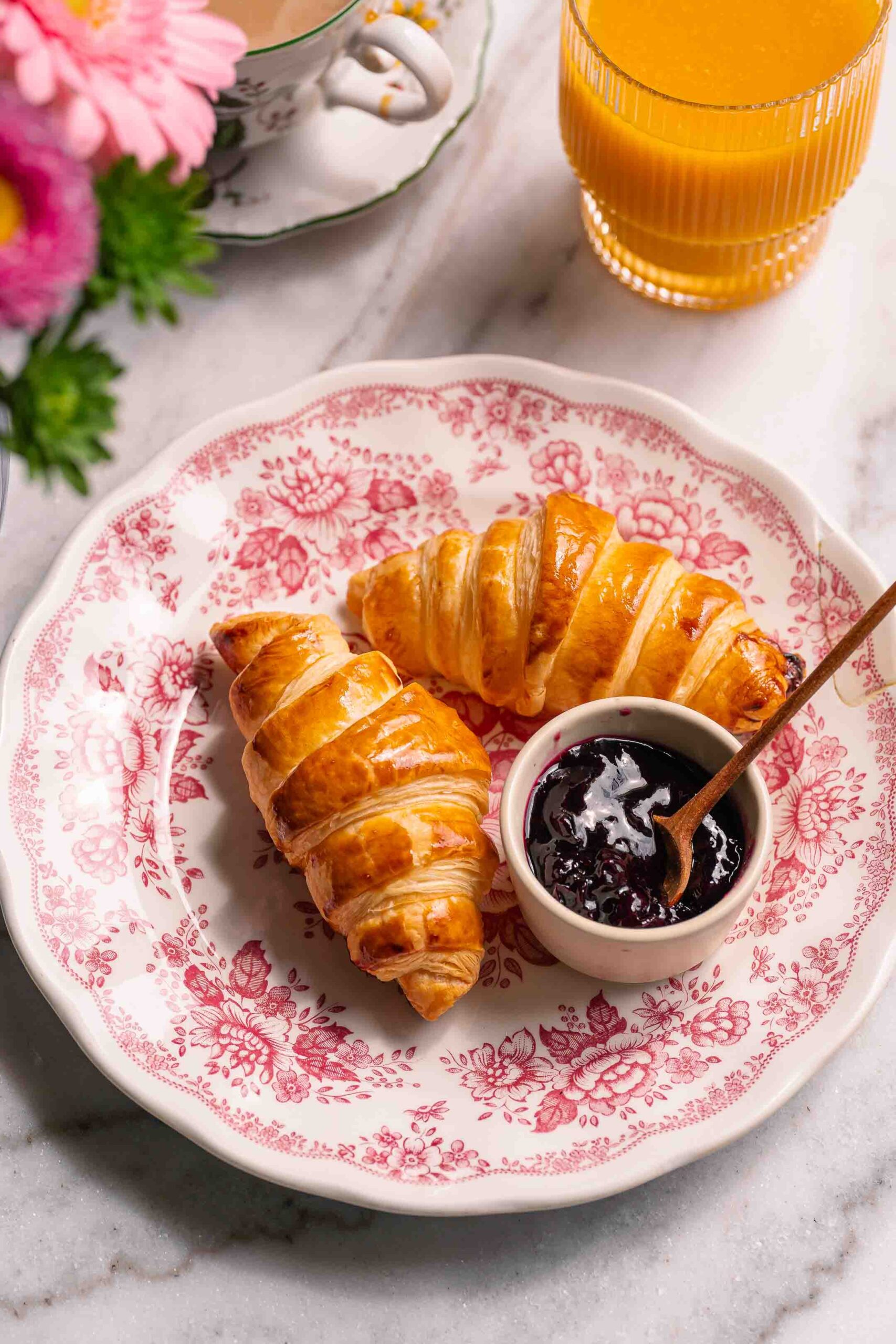 Easy Puff Pastry Croissants | Géraldine's delicacies 1 Two croissants on a decorative plate with a small bowl of dark fruit jam and a spoon, alongside a glass of orange juice and a cup of tea.