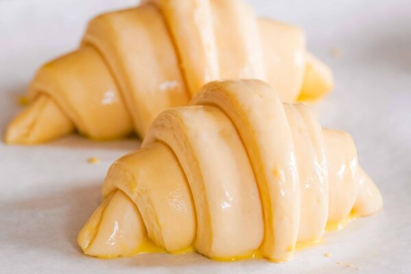 Two unbaked, egg-washed croissants are placed on a parchment-lined baking sheet.