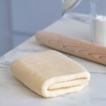 A piece of dough carefully folded on a marble countertop with a wooden rolling pin and a glass jar in the background.