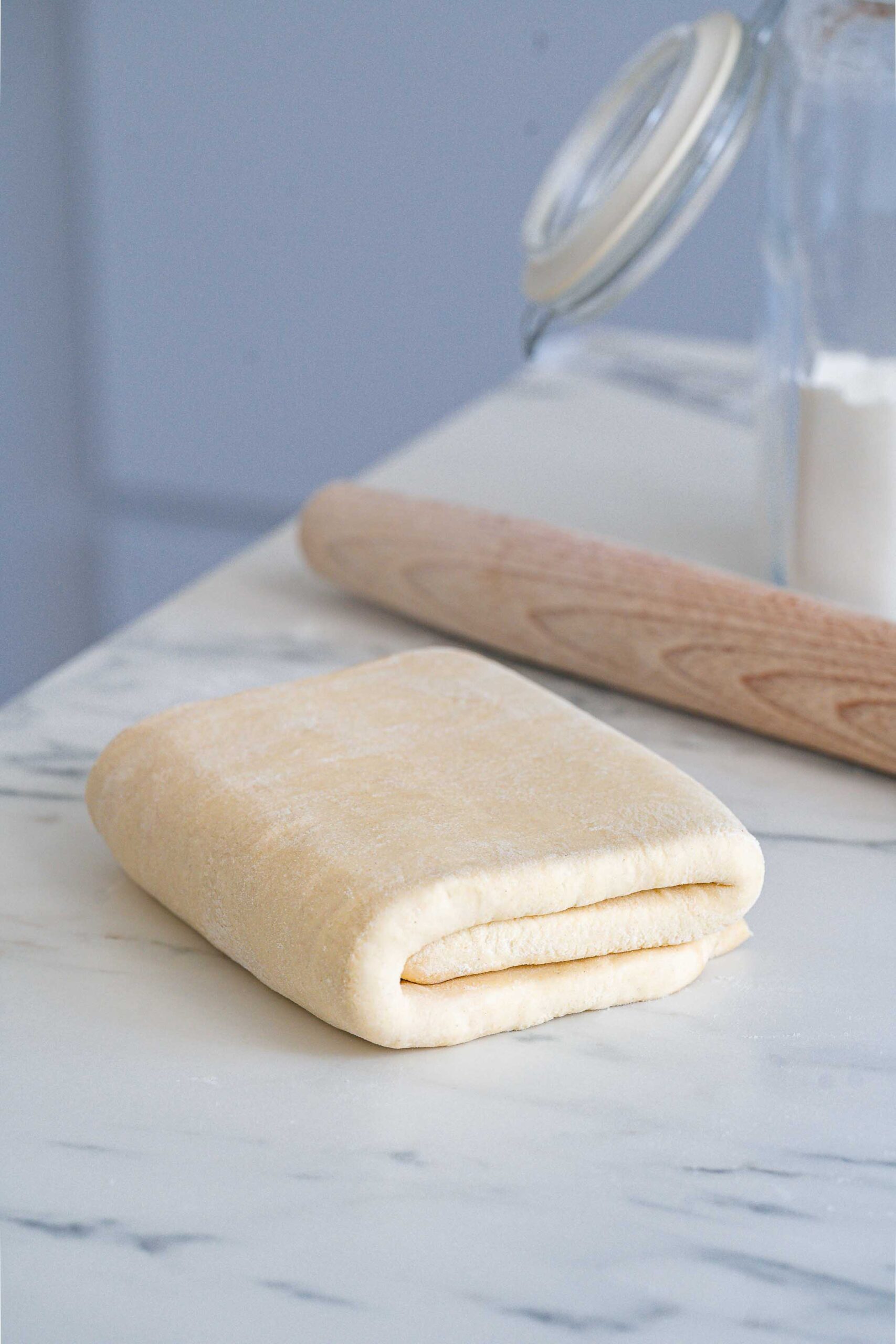 A piece of dough carefully folded on a marble countertop with a wooden rolling pin and a glass jar in the background.